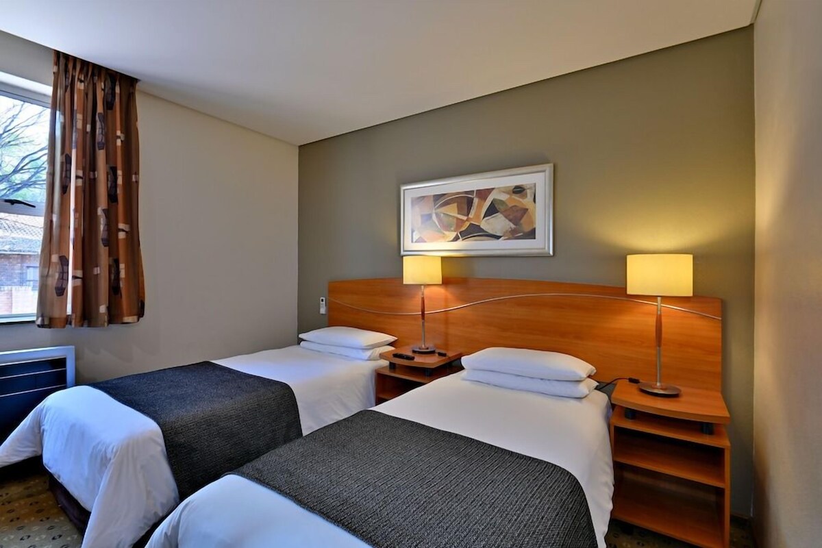 Een hotelkamer met twee bedden, een houten hoofdeinde, een wanddecoratie met abstracte kunst, en een raam met gordijnen.