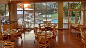 Un comedor con mesas y sillas redondas, ventanas amplias con persianas y vista a una piscina y zona de estar exterior.