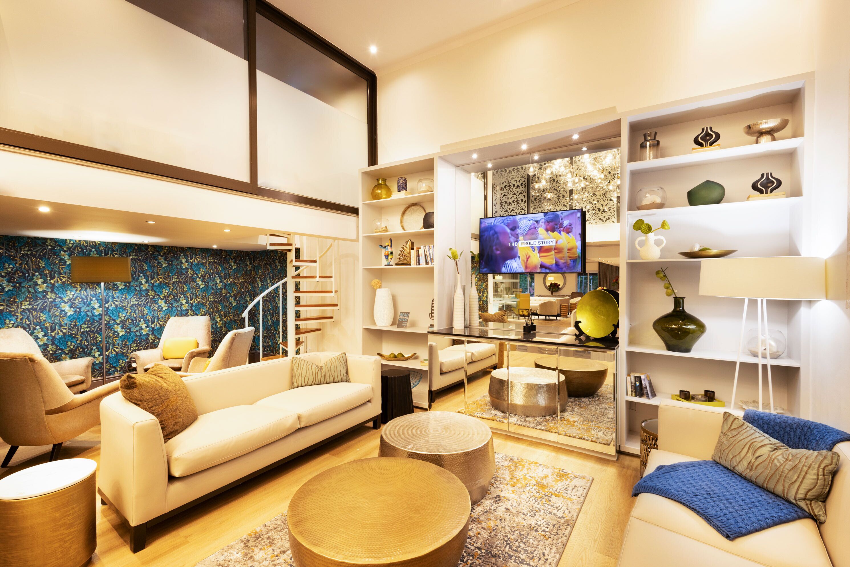 Una sala de estar moderna con un sofá, sillones, una mesa de centro y un televisor. Hay estanterías con objetos decorativos y una escalera que lleva a otro nivel.