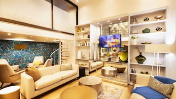 Una sala de estar moderna con un sofá, sillones, una mesa de centro y un televisor. Hay estanterías con objetos decorativos y una escalera que lleva a otro nivel.