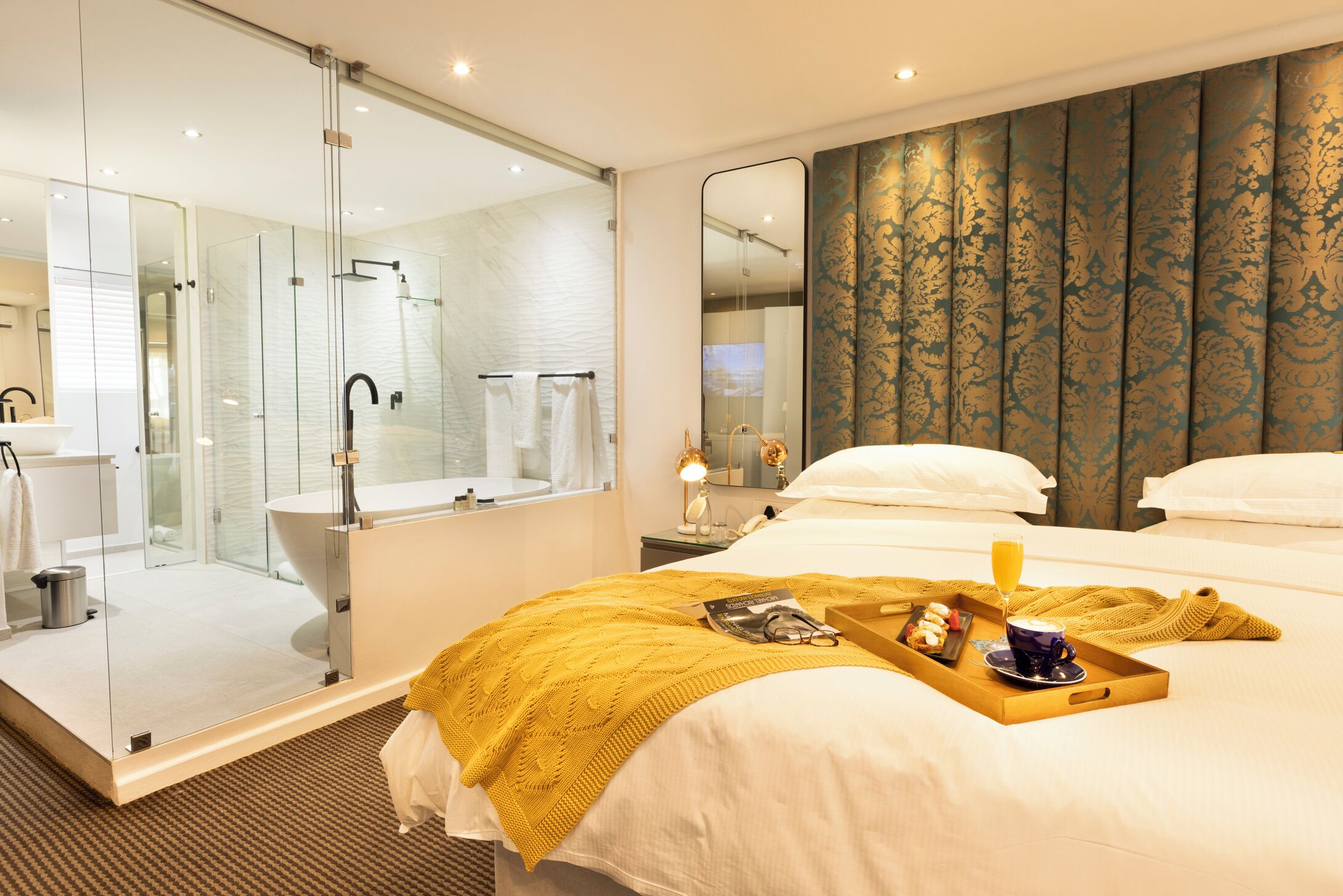 A hotel room with a glass-enclosed shower, a large bed with a yellow throw blanket, a tray with breakfast items, and a large mirror reflecting the room.