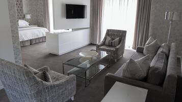 Una habitación de hotel moderna con cama, sofá, sillón, una mesa de centro de vidrio y televisor montado en la pared.