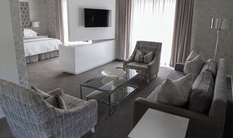 A modern hotel room with a bed, a sofa, a chair, a glass coffee table, and a TV mounted on the wall.