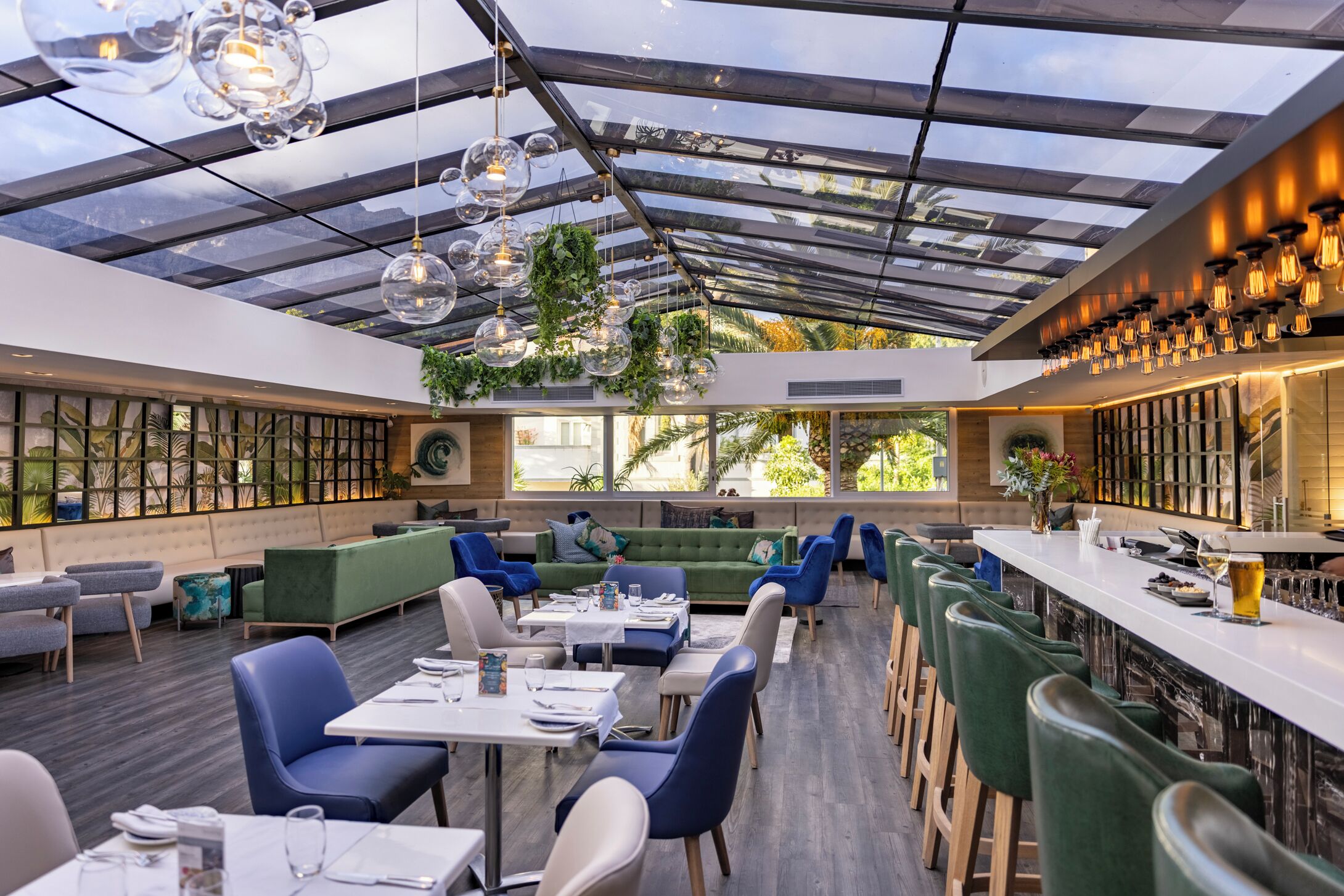 Ein modernes Restaurant mit Glasdecke, grünen Sitzgelegenheiten und einer Bar.