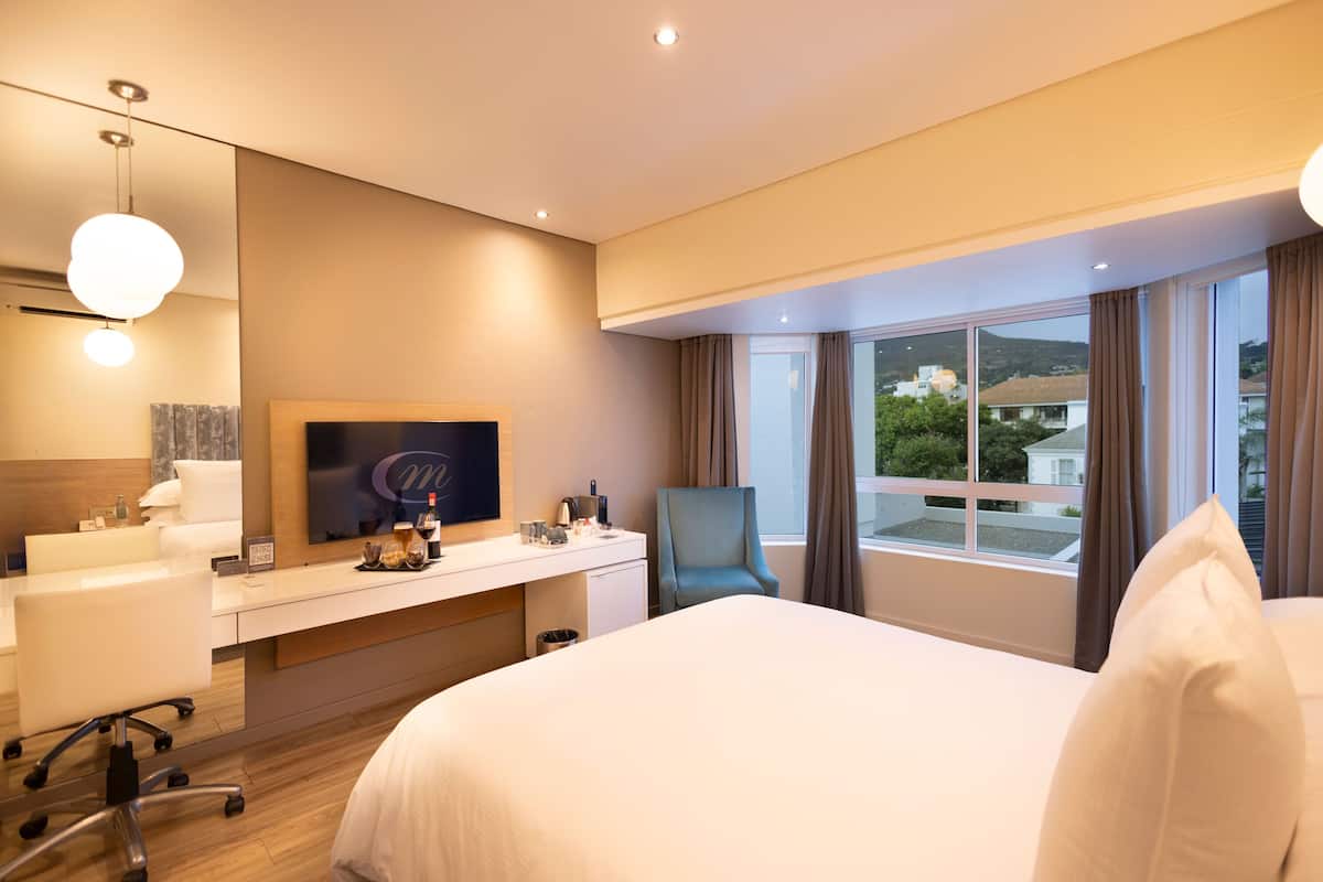 A modern hotel room with a large bed, a desk with a flat-screen TV, a chair, and a view of the city through large windows.