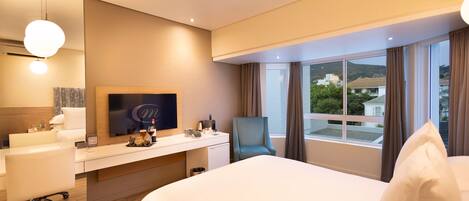 A modern hotel room with a large bed, a desk with a flat-screen TV, a chair, and a view of the city through large windows.