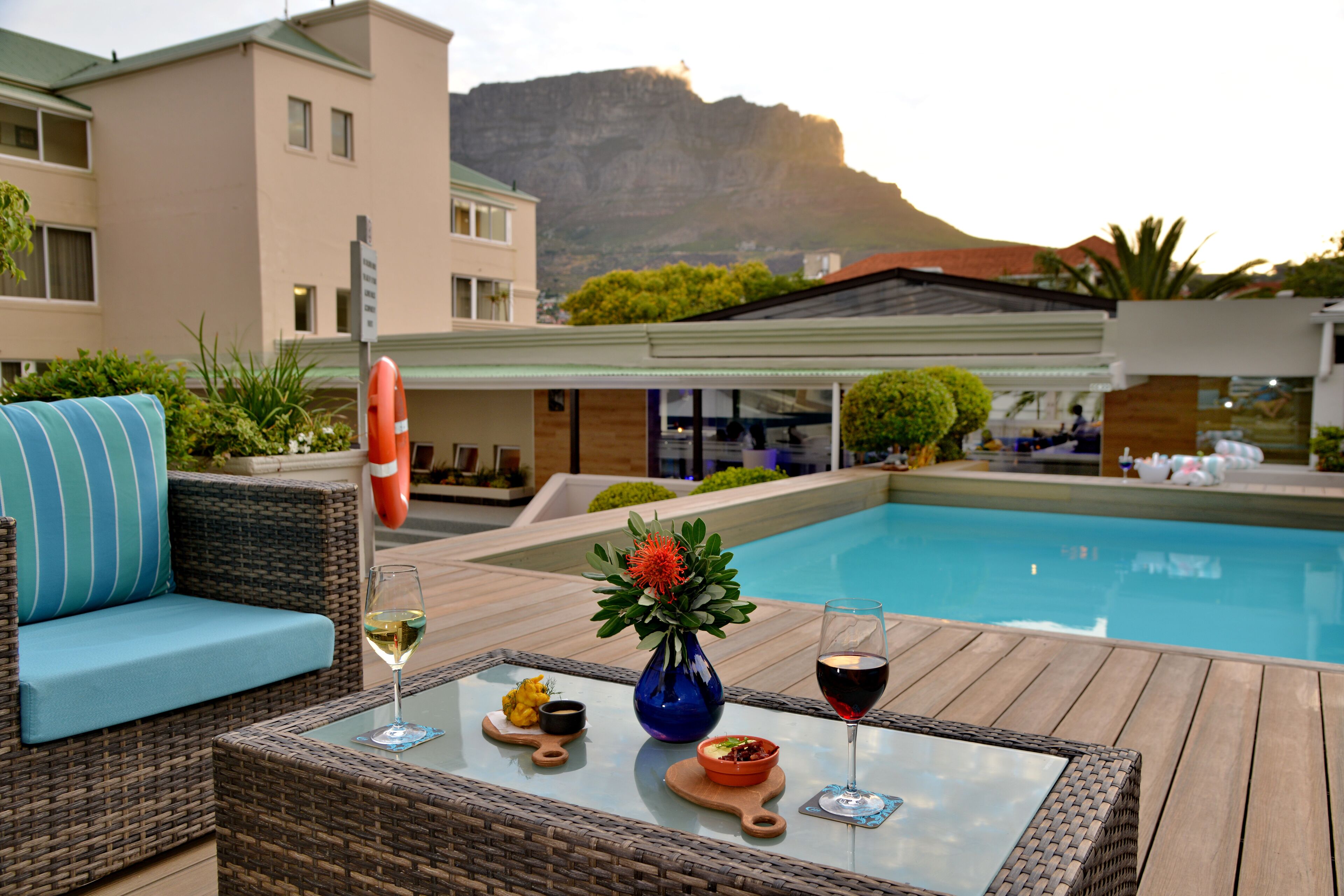A poolside area with a wicker table, a glass of white wine, a glass of red wine, a bowl of fruit, and a vase with a flower.