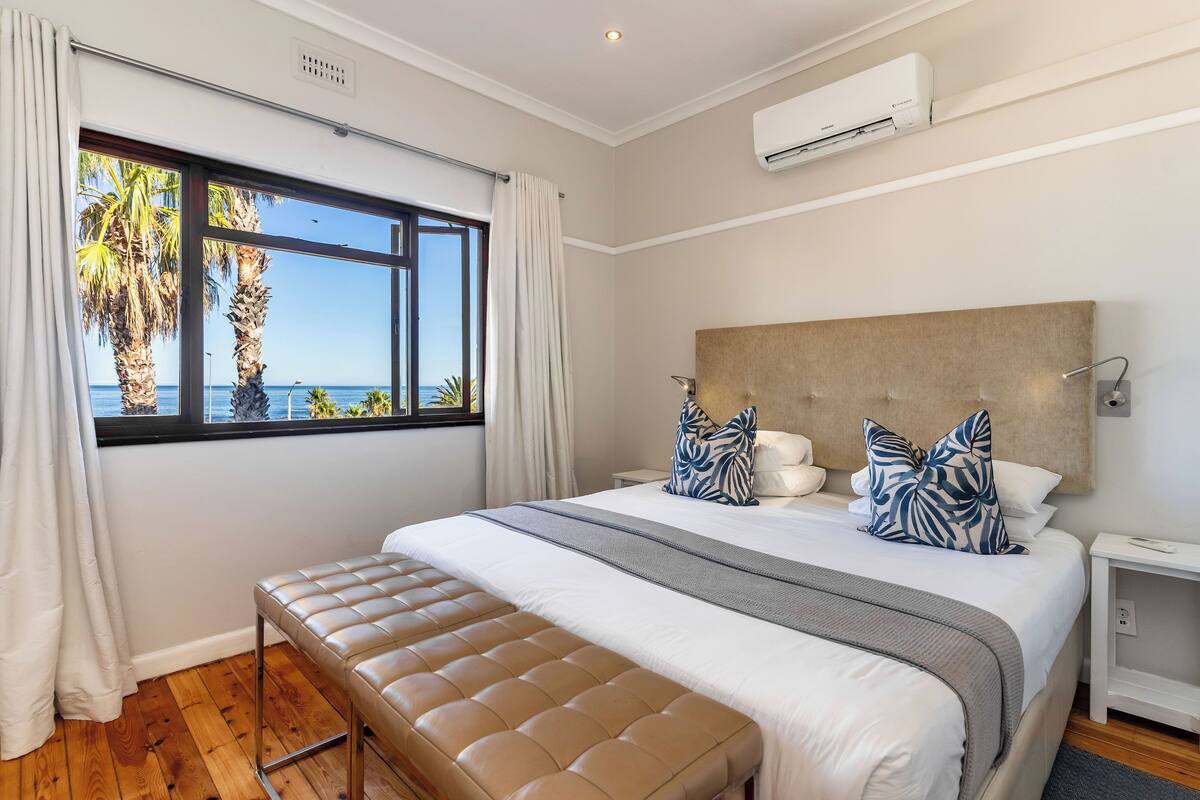 A bedroom with a large bed, a bench, a nightstand, and a view of the sea through a window.