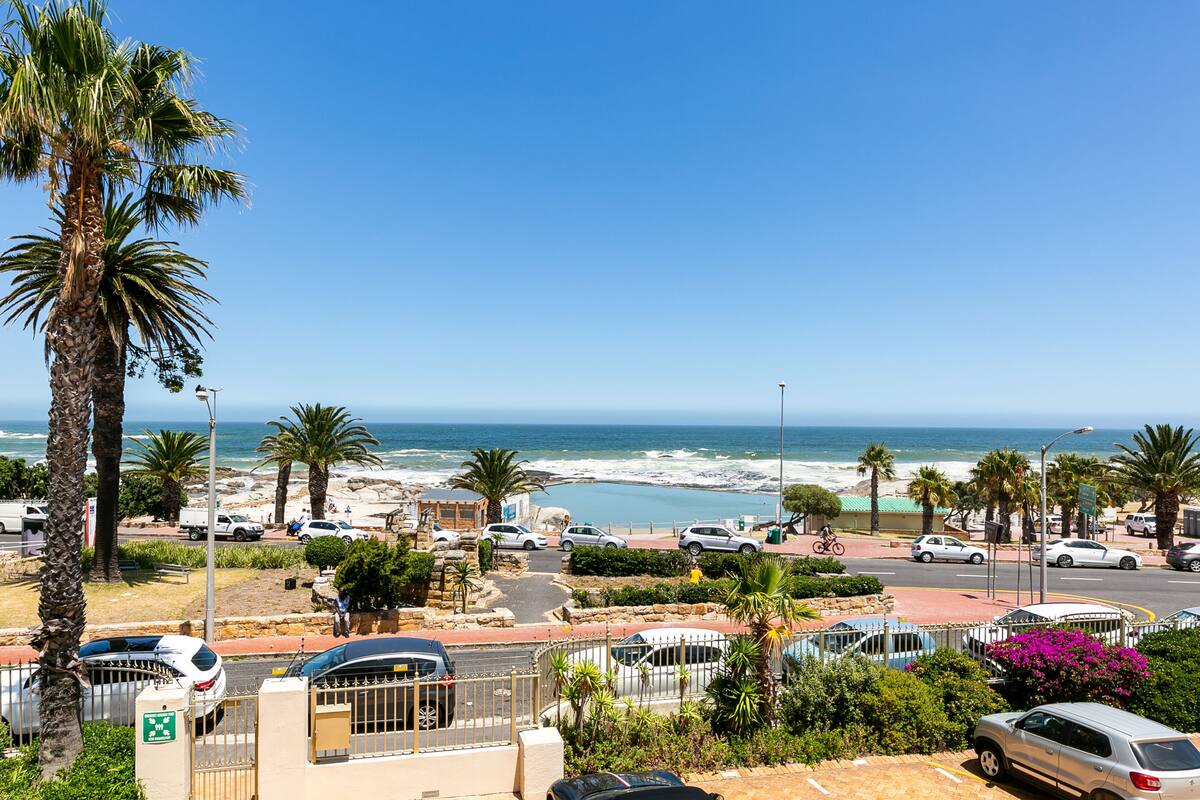 A coastal street with parked cars, palm trees, and a view of the beach and ocean.