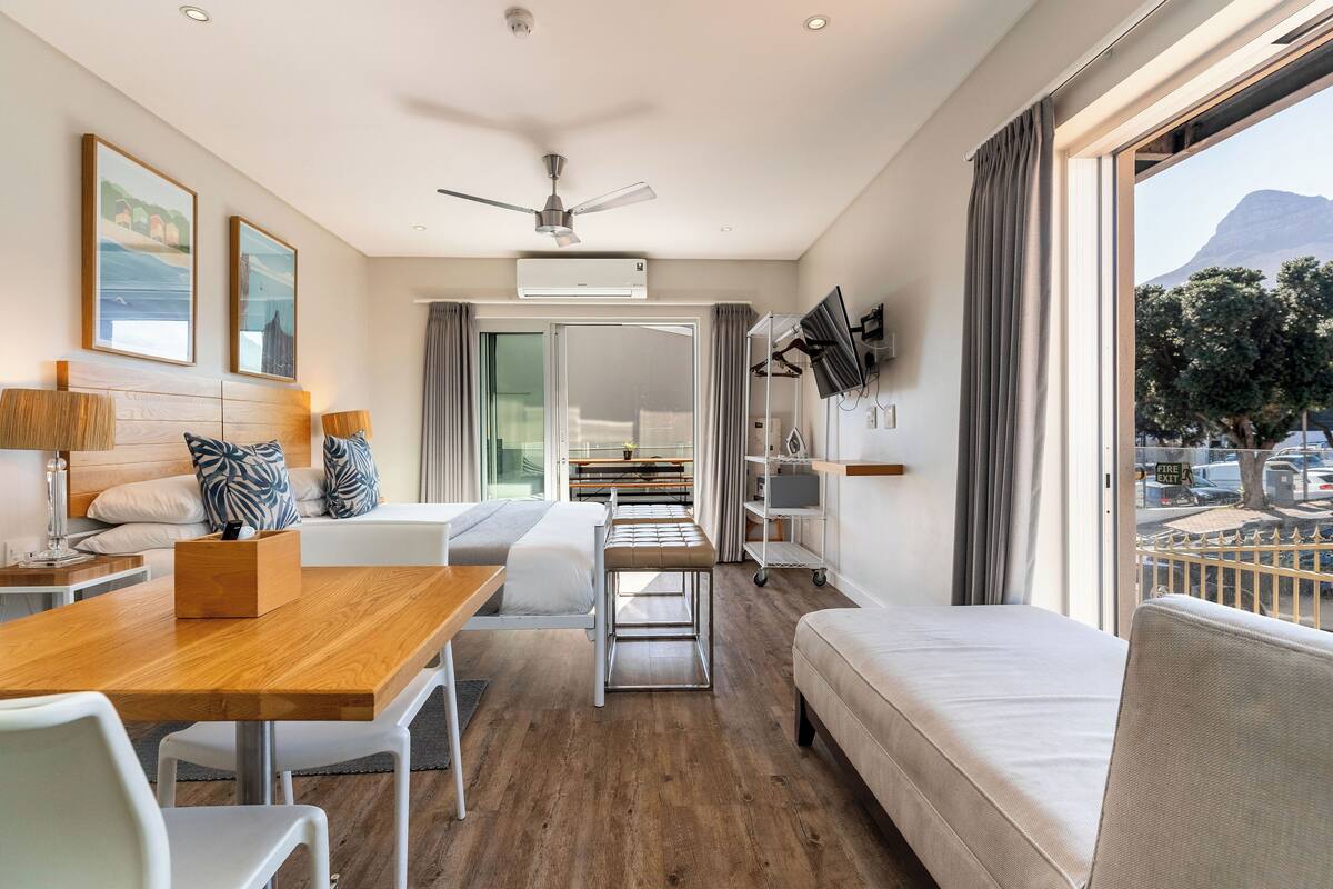 A modern hotel room with a wooden dining table, a sofa, a bench, and a TV mounted on the wall.