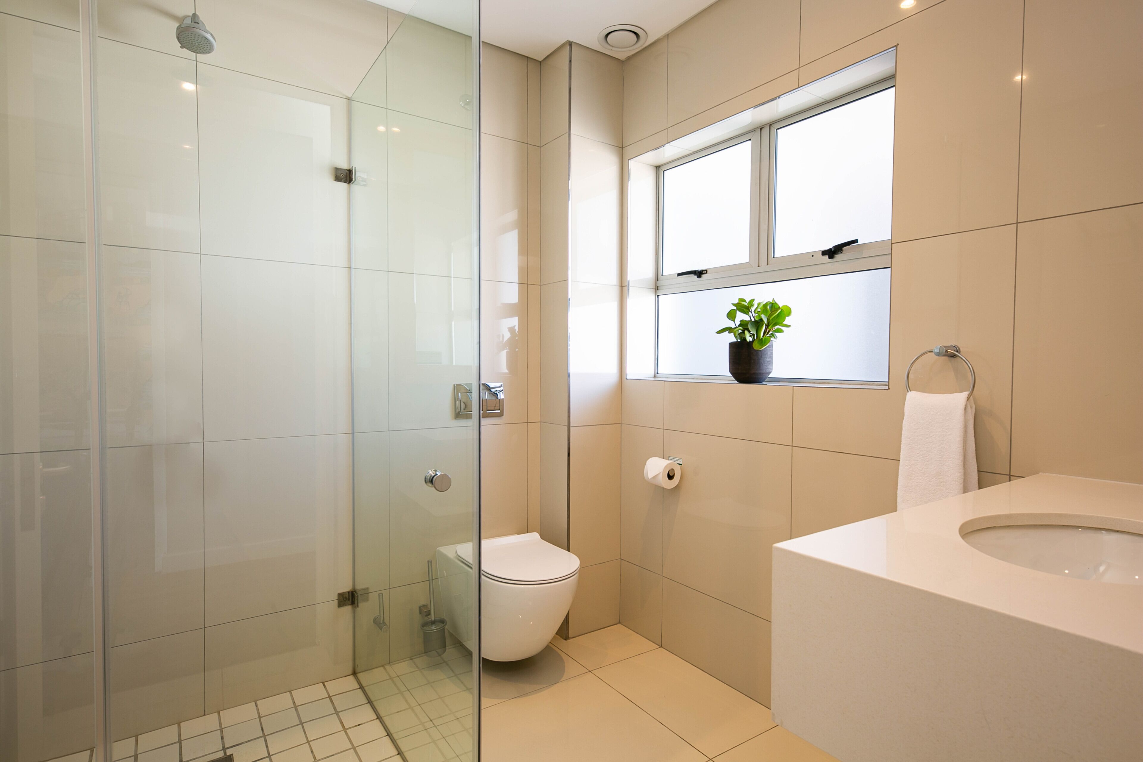 Een moderne badkamer met een glazen douchecabine, toilet, wastafel met spiegel en raam met plant.