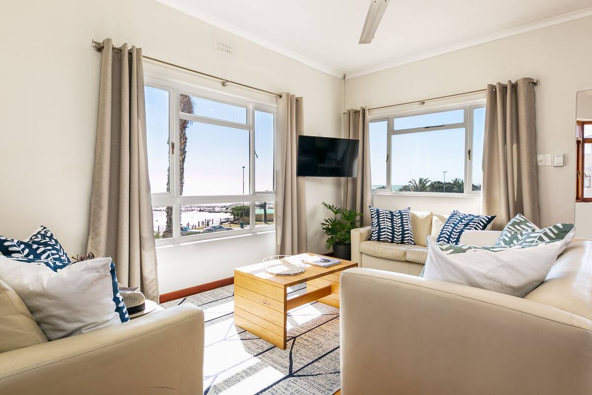 A well-lit living room with two sofas, a coffee table, and a flat-screen TV mounted on the wall. Large windows with curtains provide natural light and views of the outdoors.