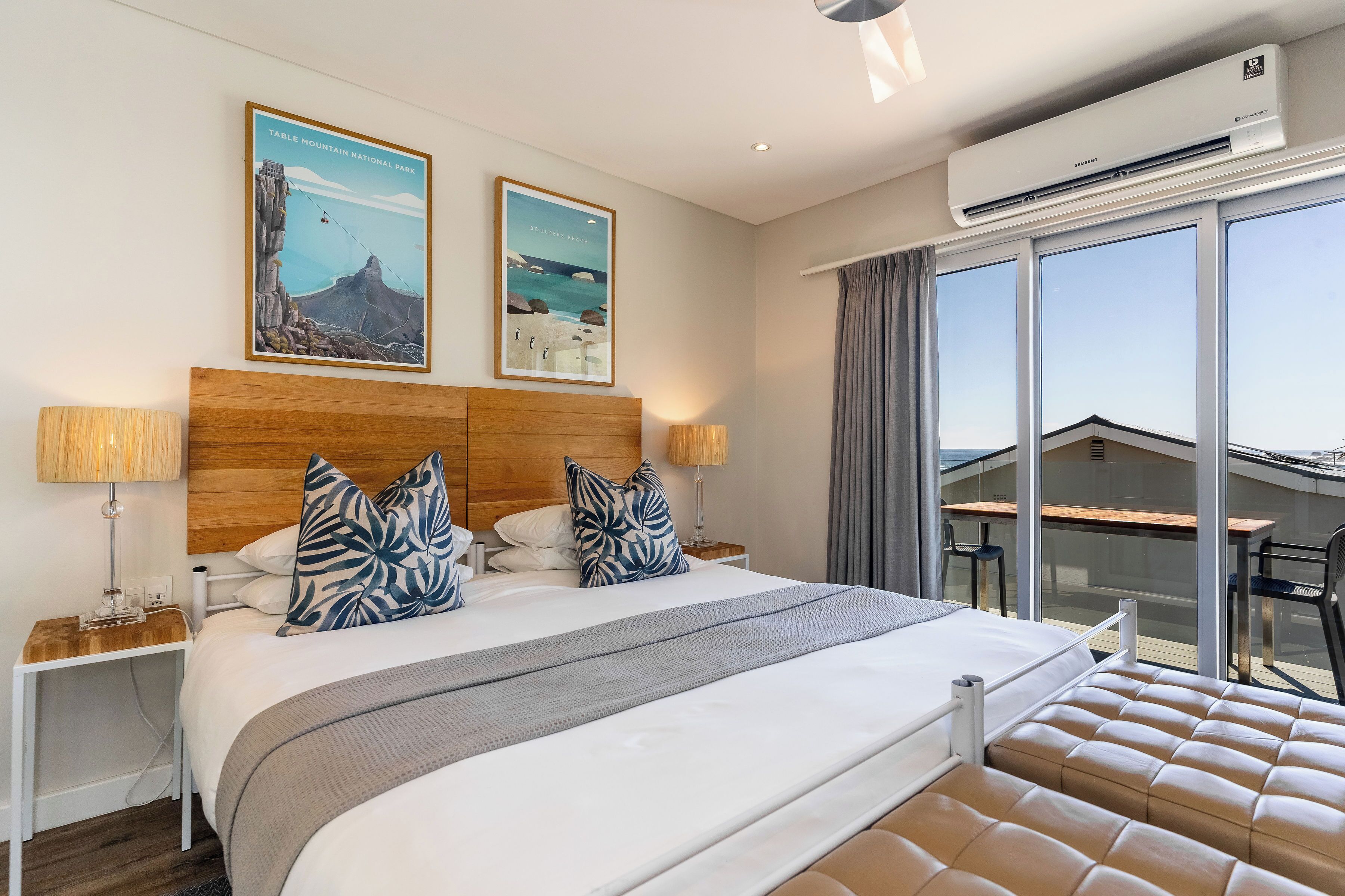A hotel room with a large bed, two posters on the wall, an air conditioning unit, a balcony with a table and chairs, and a view of the mountains.