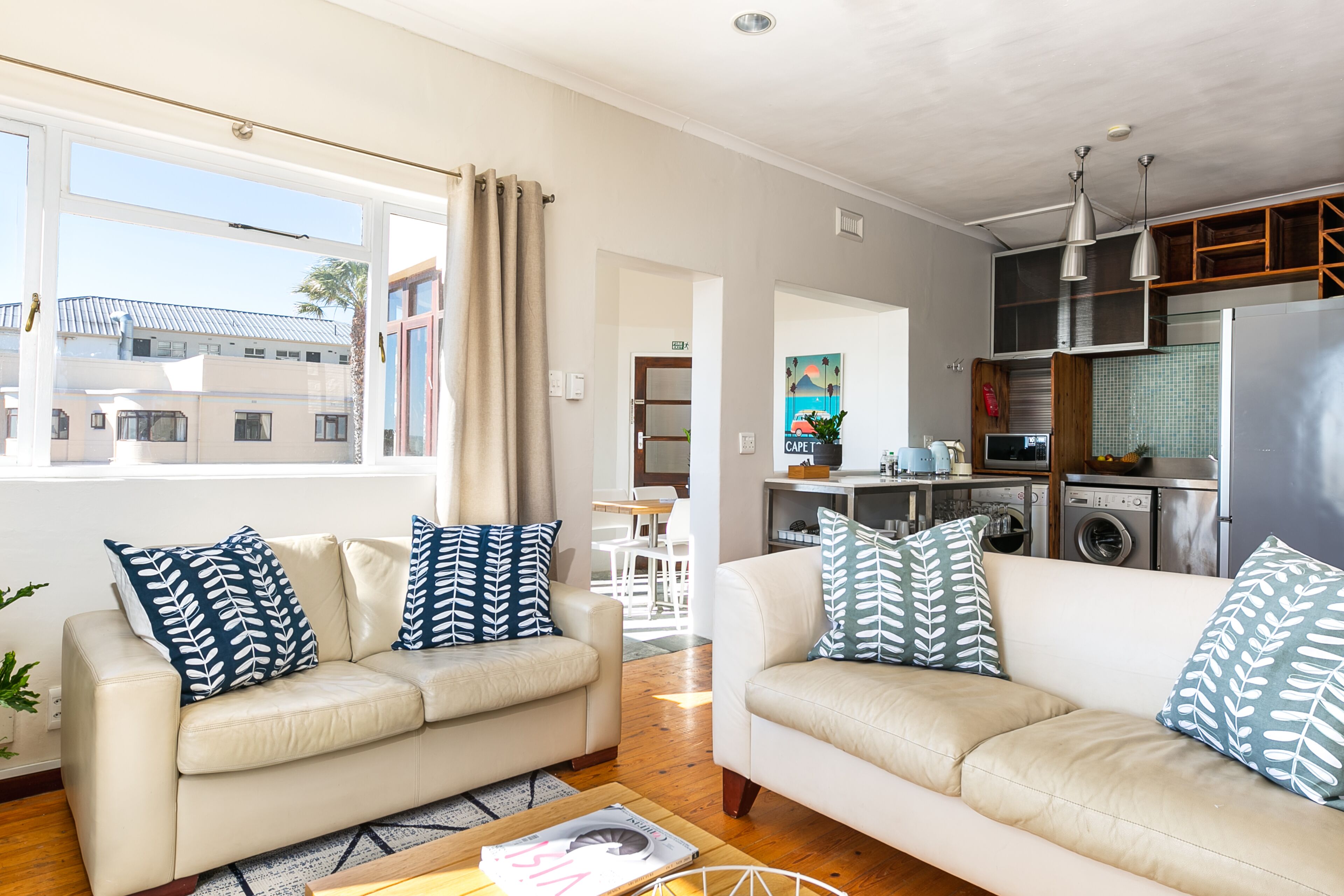 A modern living room with two beige sofas, patterned throw pillows, and a coffee table. A kitchen with stainless steel appliances and wooden cabinets is visible in the background.