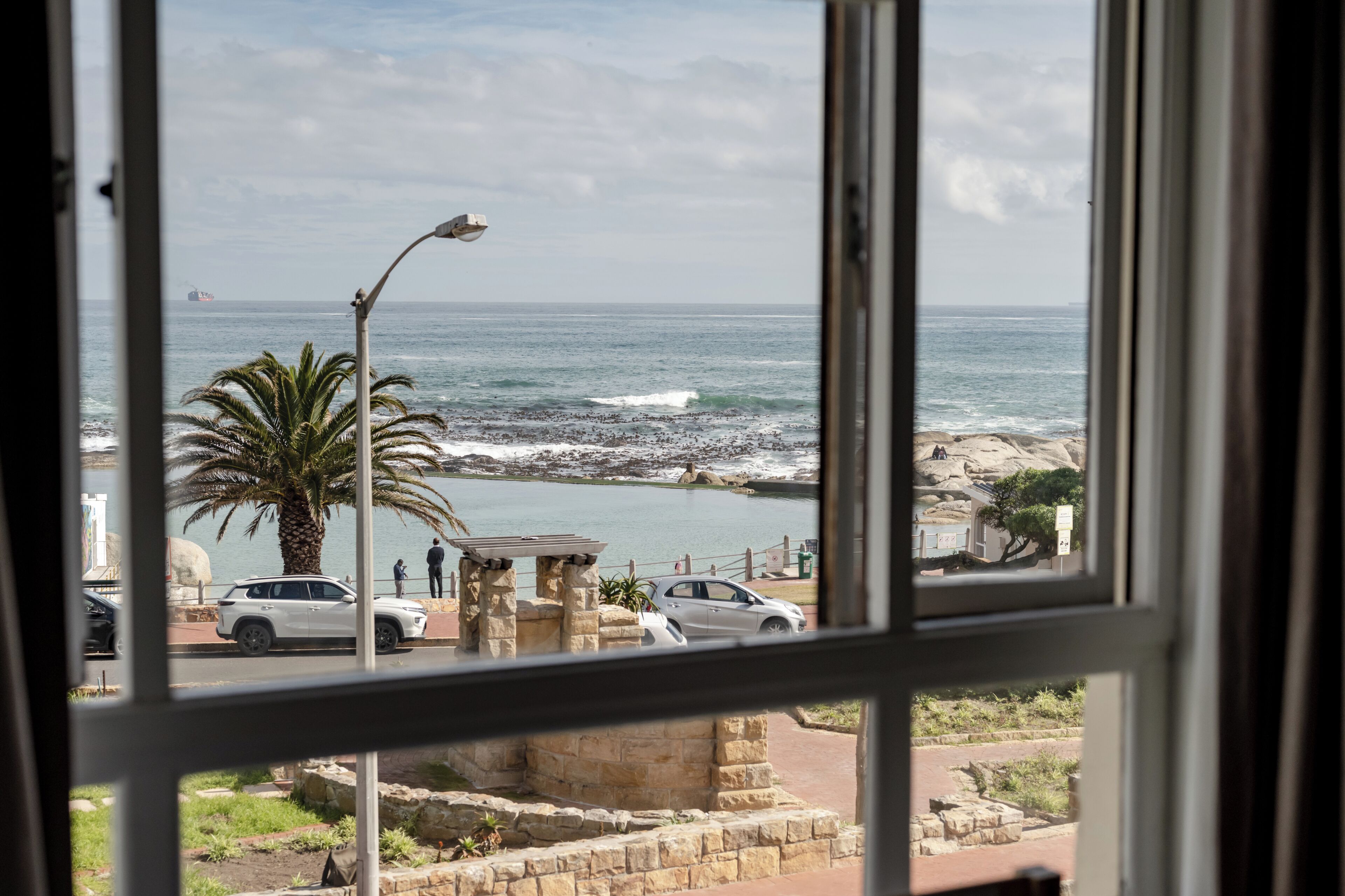 A coastal view through a window, including a palm tree, a street lamp, and a rocky shoreline.