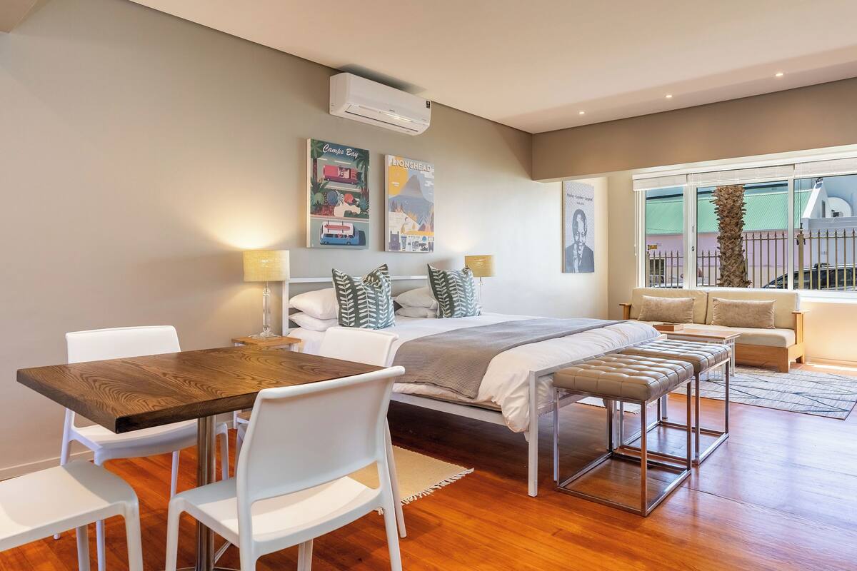 A modern bedroom with a wooden dining table, white chairs, a bed with a grey blanket, and a large window with a view of a palm tree.