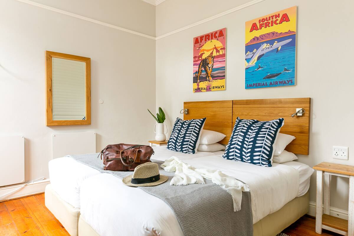 A neatly made bed with a wooden headboard, a brown leather bag, a straw hat, and a white blanket. There are two pillows with leaf patterns, a small white vase with a plant, and a framed picture of a leopard on the wall.