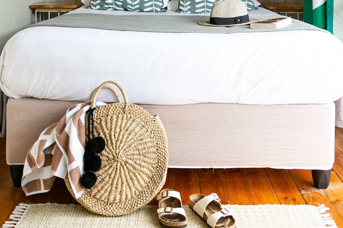 A neatly made bed with a striped pillow, a woven bag, and sandals on the floor. Two posters of Africa are on the wall.