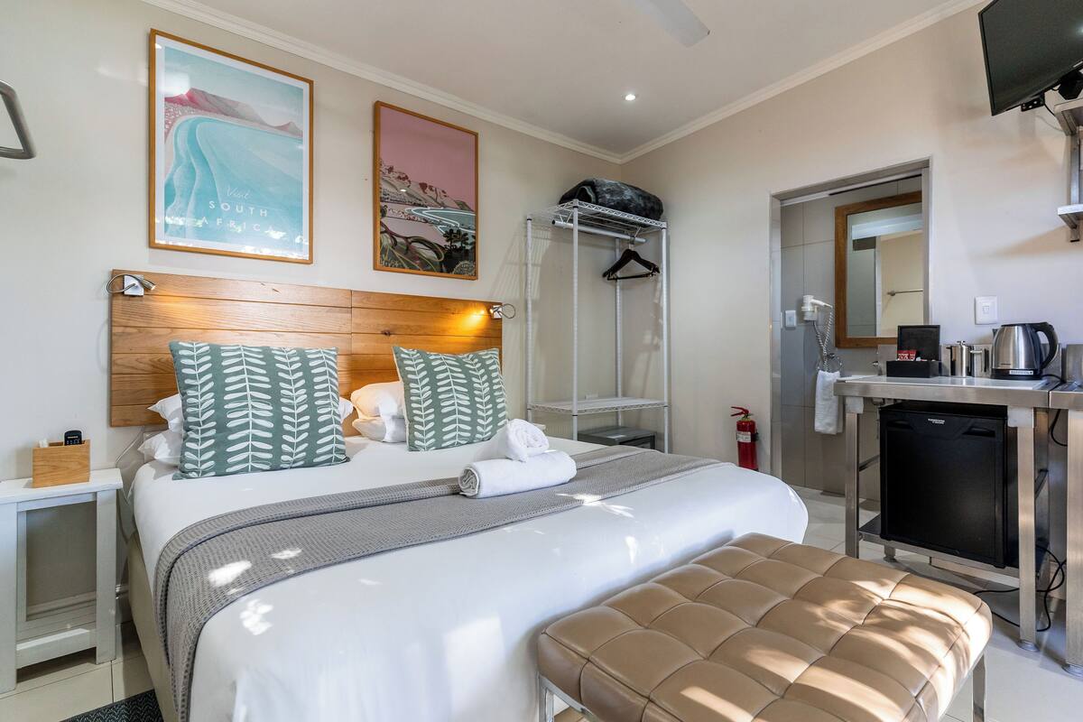 Habitación de hotel con cama, un banco, escritorio con cafetera, espejo y armario.