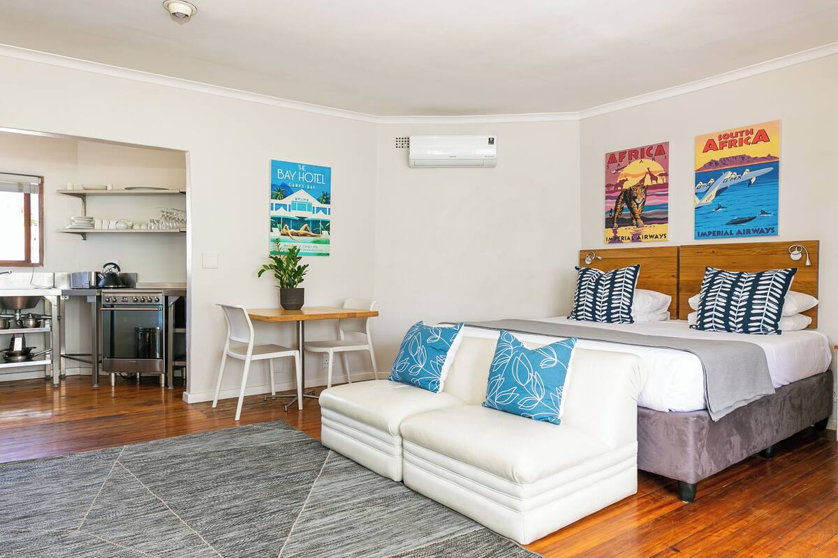 A modern hotel room with a bed, a sofa, a dining table, and a kitchenette.