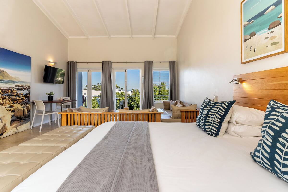 Una habitación de hotel moderna con una cama grande, zona de estar, televisor y vistas al exterior.