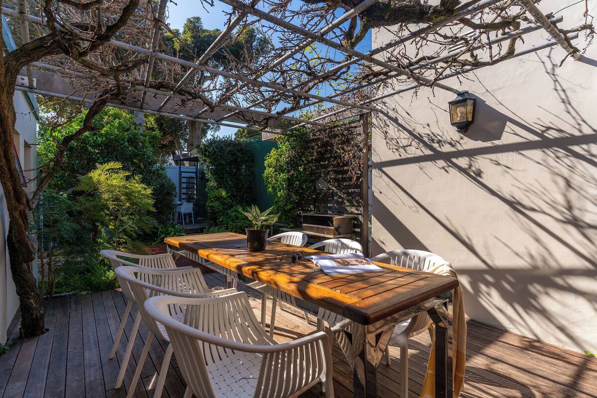 An outdoor patio with a wooden table and white chairs, surrounded by greenery and a pergola structure.