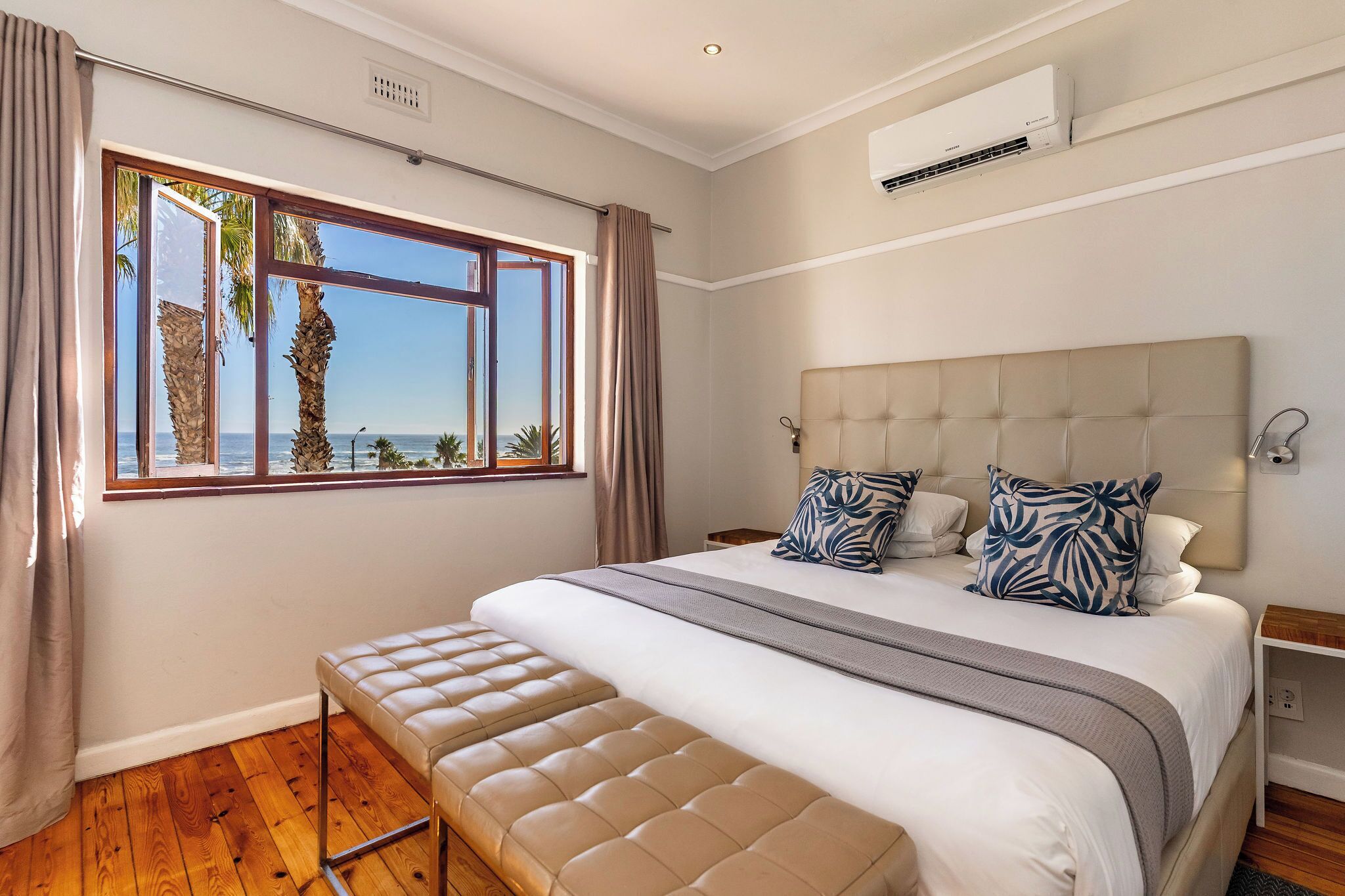 A bedroom with a large bed, a bench, a window with curtains, an air conditioning unit, and a view of palm trees and the sea.
