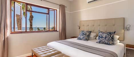 Un dormitorio con una cama grande, una banca, una ventana con cortinas, un equipo de aire acondicionado y vistas a palmeras y al mar.