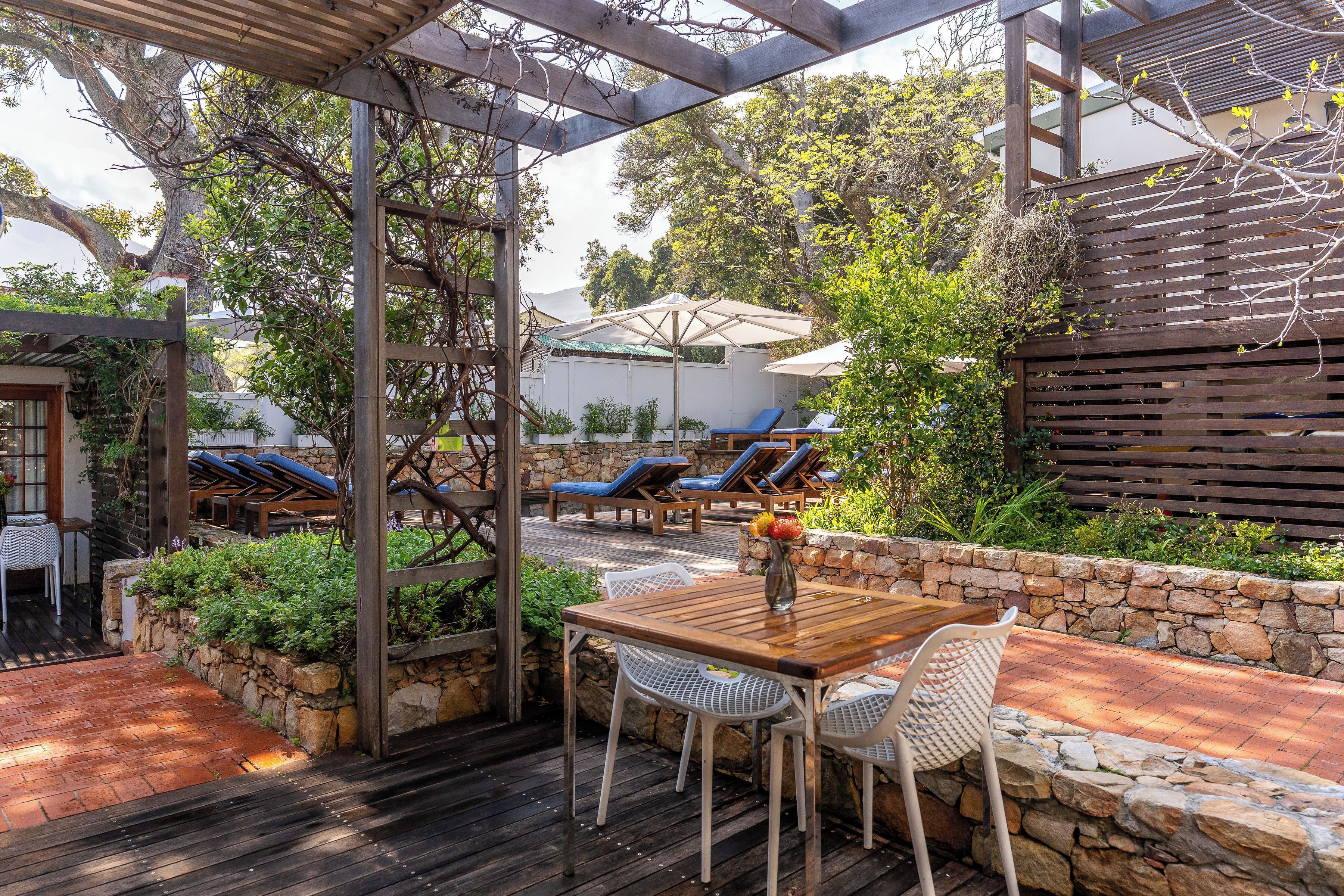 An outdoor patio with a wooden table and chairs, surrounded by stone walls and greenery.