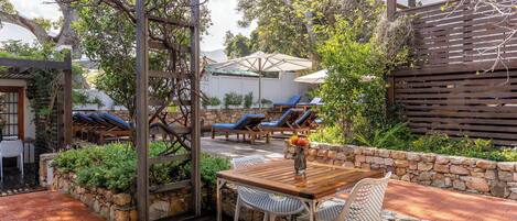 An outdoor patio with a wooden table and chairs, surrounded by stone walls and greenery.