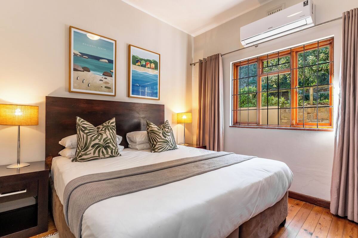 A neatly made bed with a headboard, two wall-mounted air conditioning units, a window with curtains, and two framed pictures on the wall.