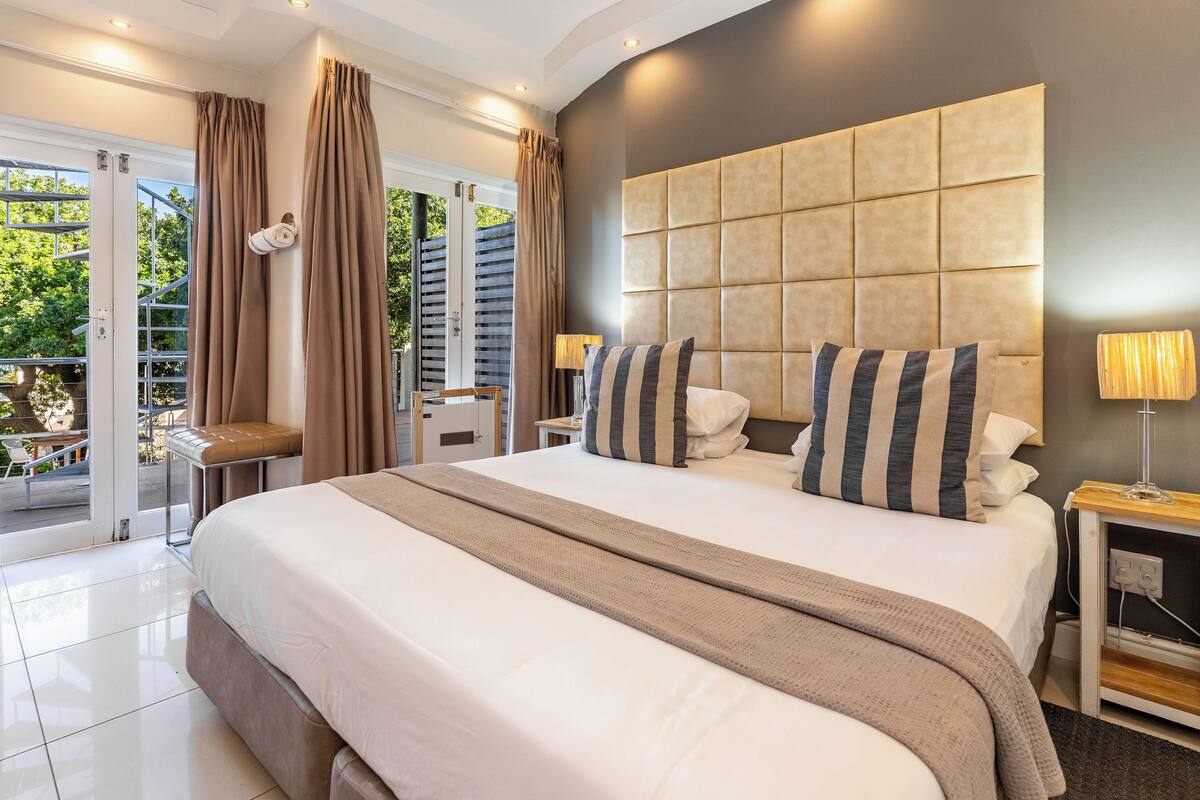 A modern hotel room with a large bed, a headboard, a bedside table, and a view of the outdoors.
