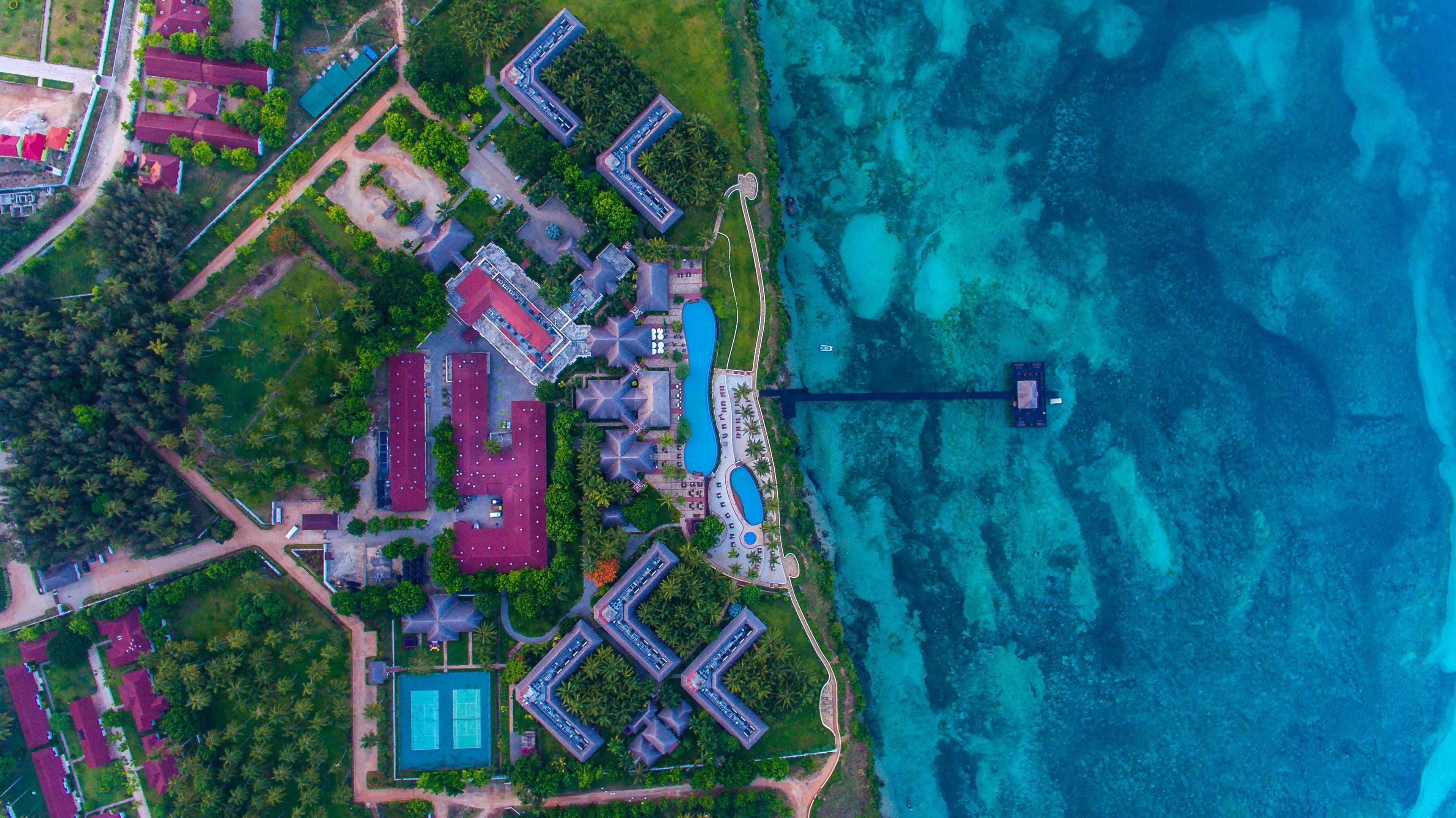 Une vue aérienne d’un complexe hôtelier comprenant plusieurs bâtiments, une grande piscine et une jetée s’avançant dans l’océan.