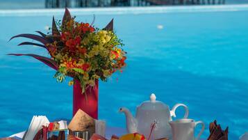 A breakfast table with fruits, a teapot, and a vase of flowers by a pool with a view of a pier.