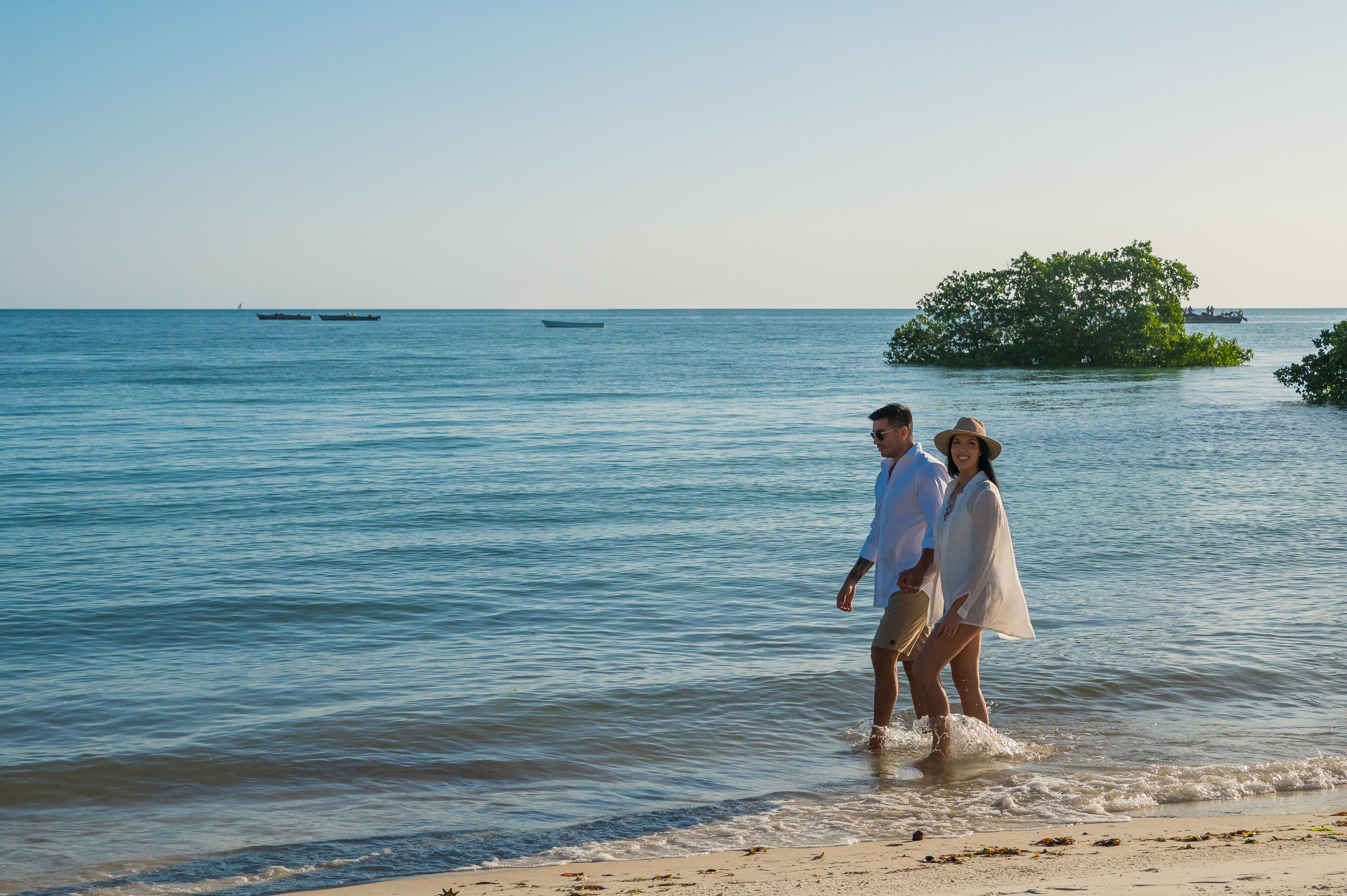 Una pareja caminando en una playa con aguas tranquilas y barcos a lo lejos.