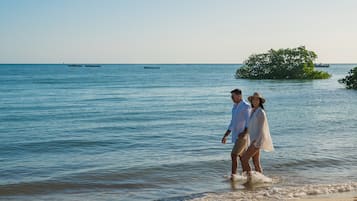 Una pareja caminando en una playa con aguas tranquilas y barcos a lo lejos.