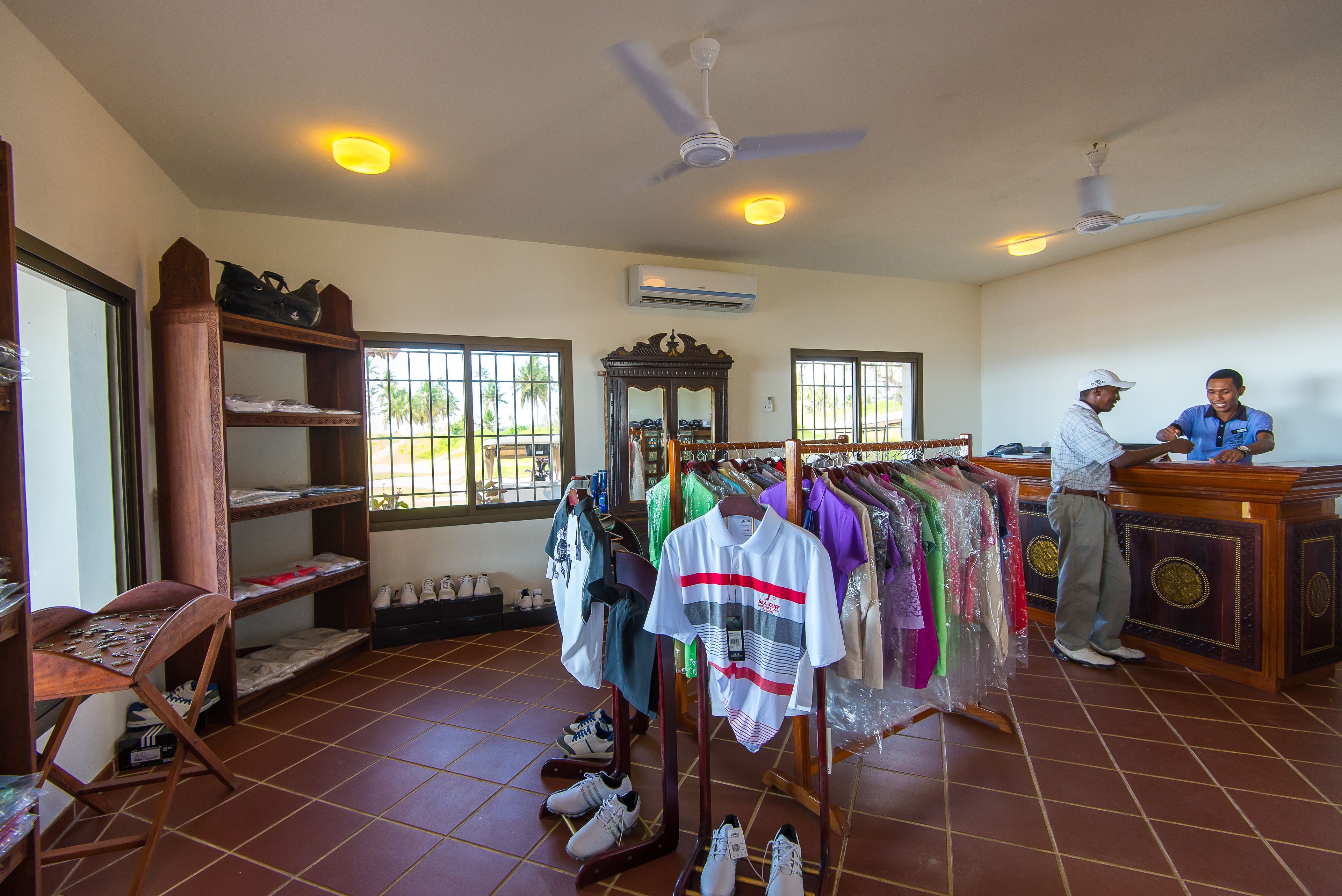 A room with a wooden desk, shelves with shoes, a clothing rack, and a ceiling fan.