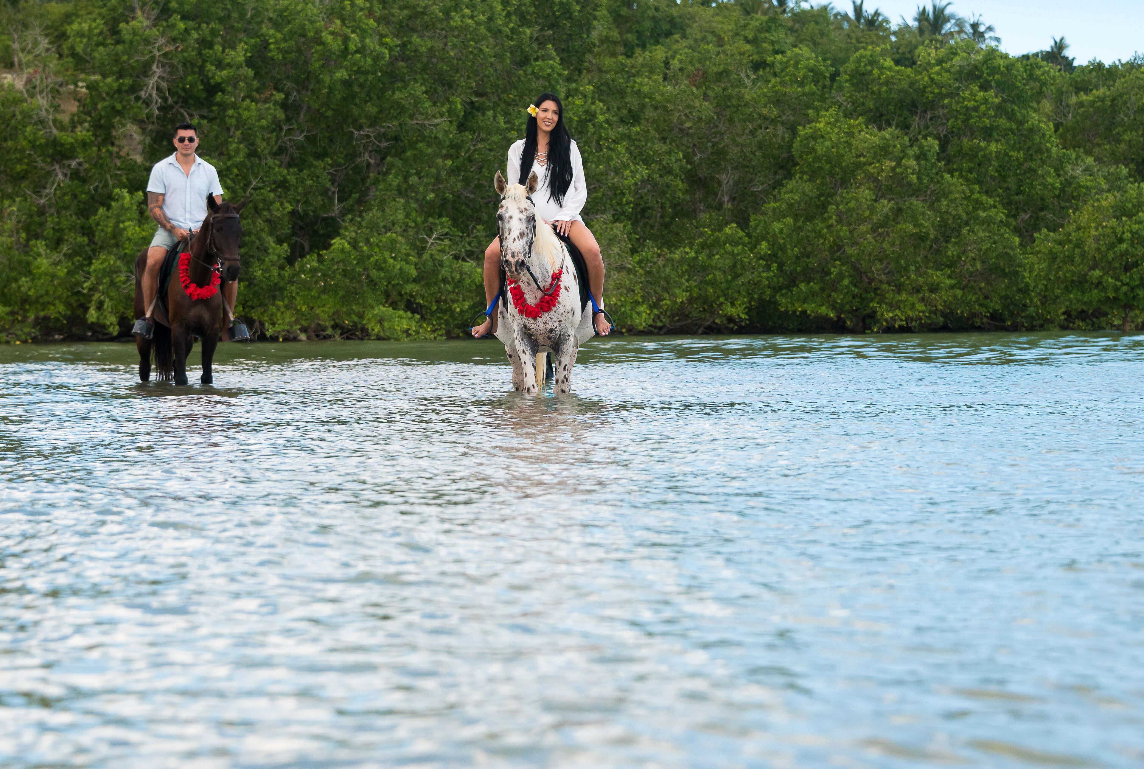 Two people riding horses through a body of water with dense greenery in the background.