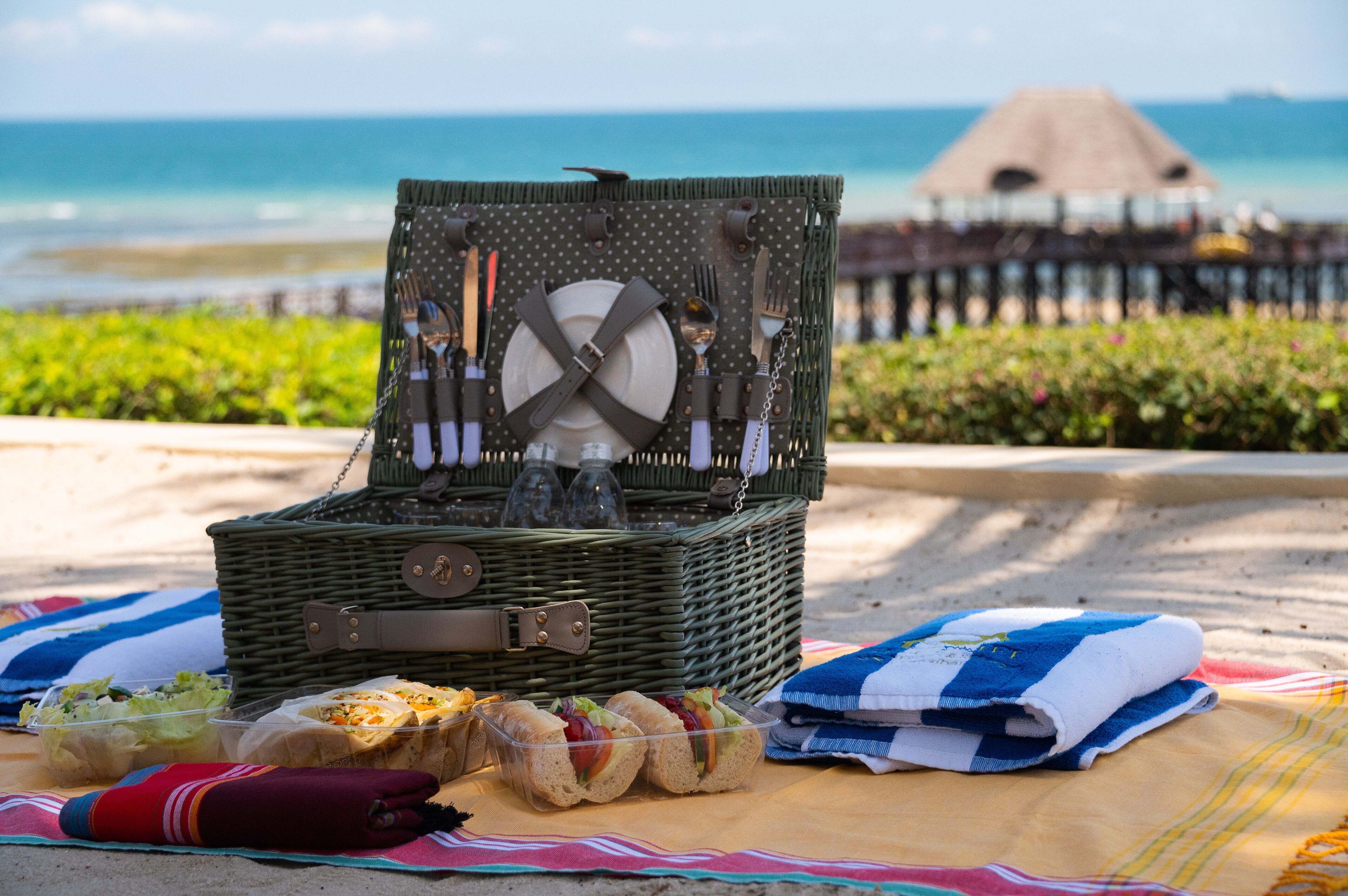 Montaje de picnic en la playa, con una cesta de mimbre que contiene utensilios, una toalla doblada y sándwiches sobre una manta.