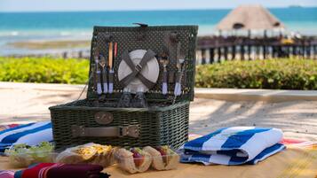 Montaje de picnic en la playa, con una cesta de mimbre que contiene utensilios, una toalla doblada y sándwiches sobre una manta.