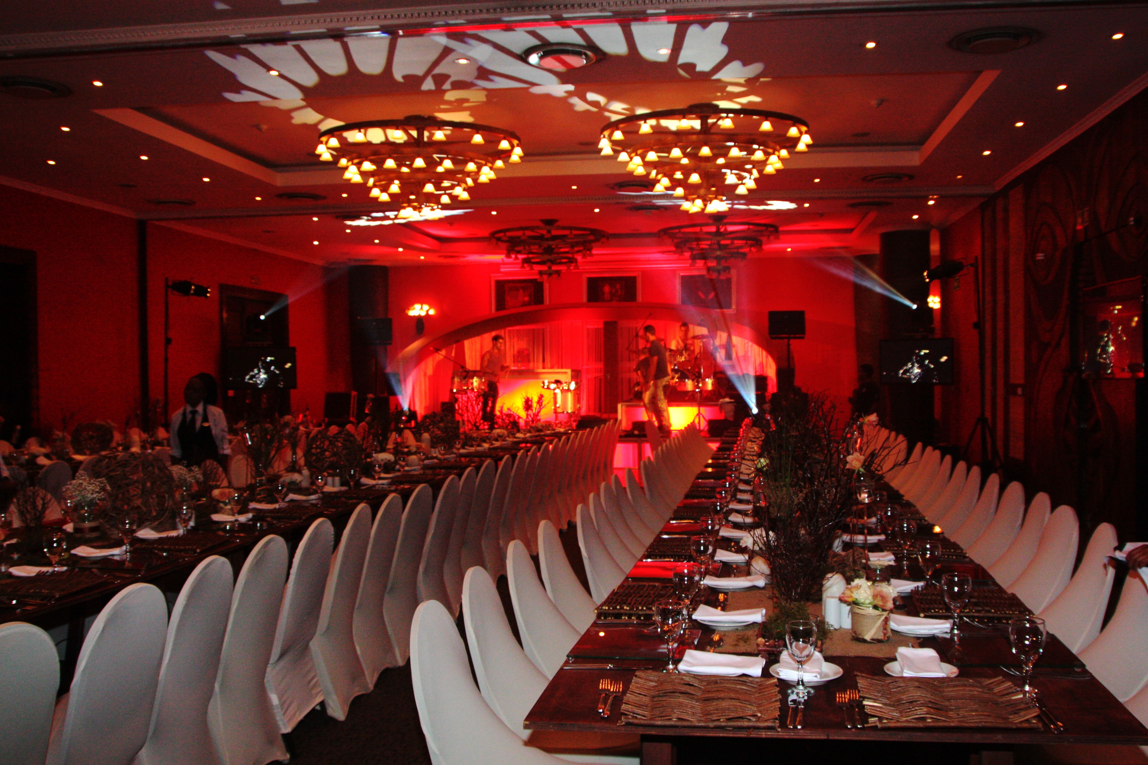 A long dining table with white chairs, set for a formal meal in a dimly lit room with red ambient lighting and chandeliers.
