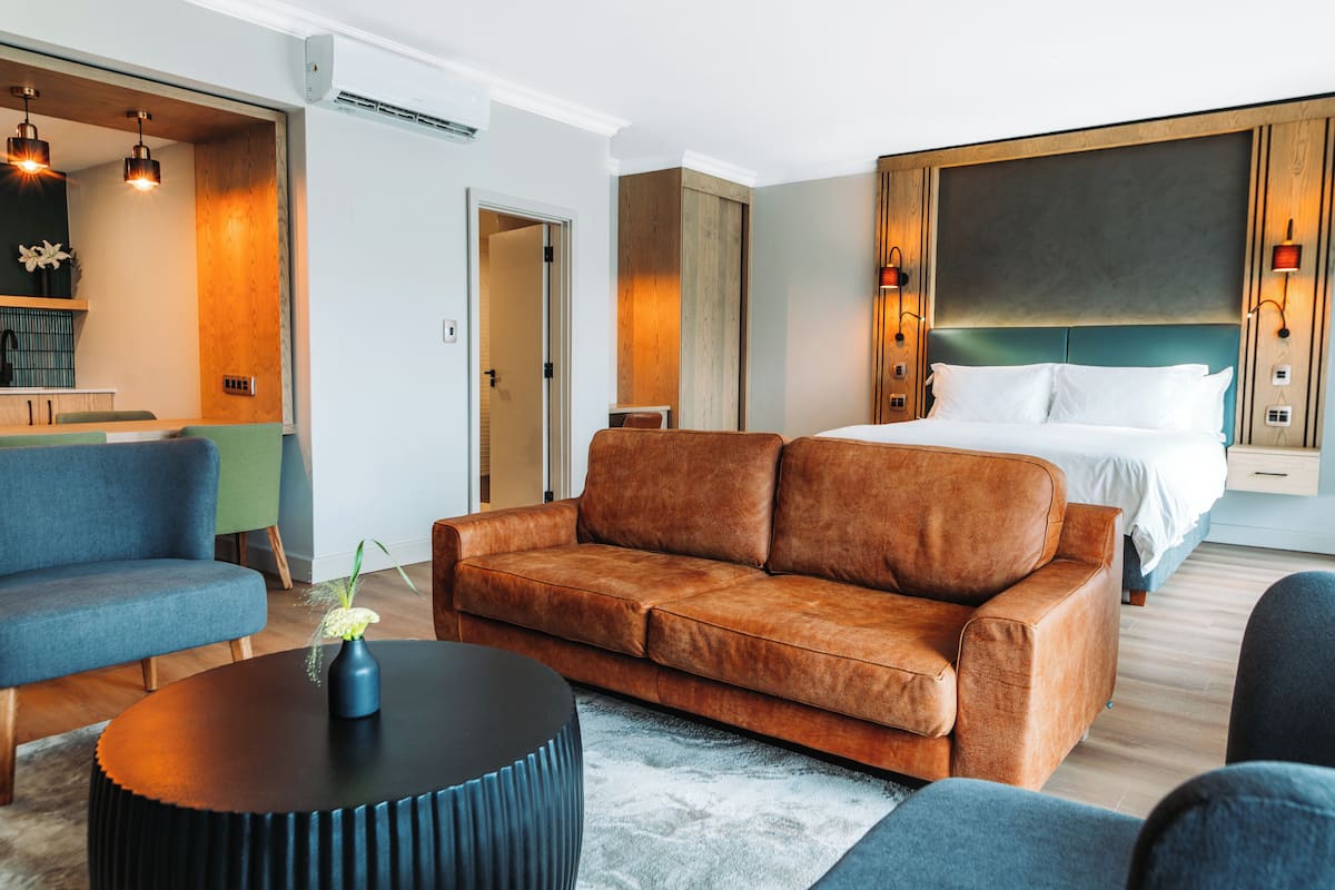 A modern hotel room with a brown leather sofa, a round black coffee table, and a bed with white linens.