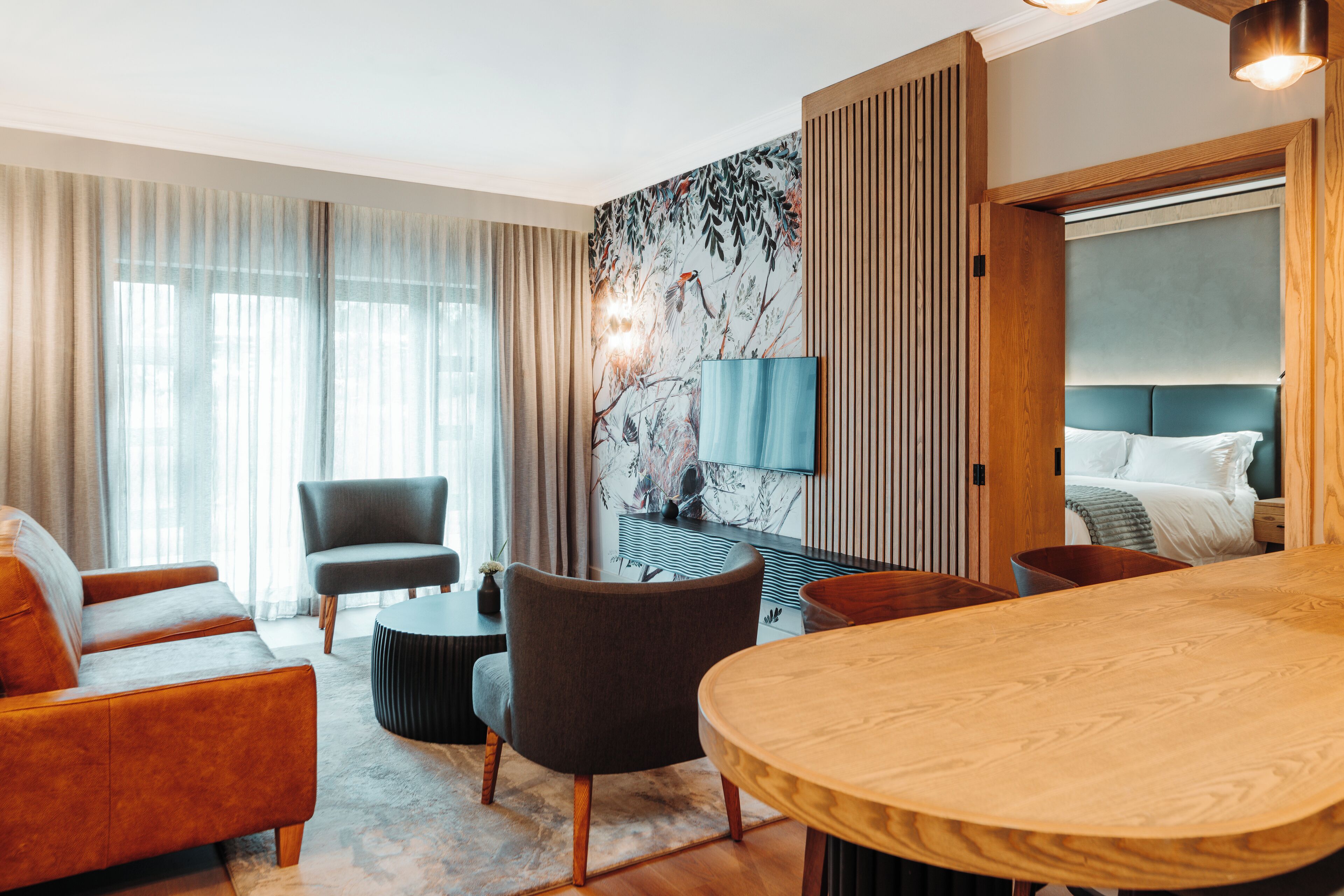 A modern hotel room with a wooden dining table, a sofa, a chair, a TV, and a bed with a headboard.