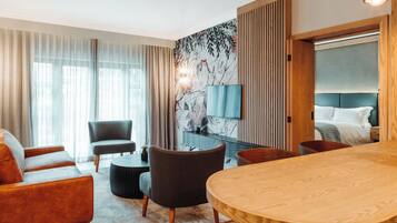 A modern hotel room with a wooden dining table, a sofa, a chair, a TV, and a bed with a headboard.