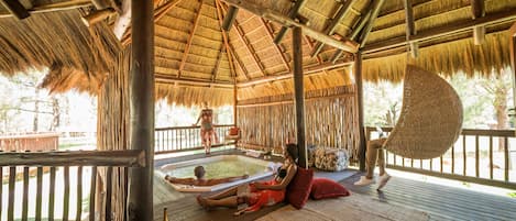 A wooden deck with a thatched roof, a hot tub, and a lounge area with a woven chair and cushions.