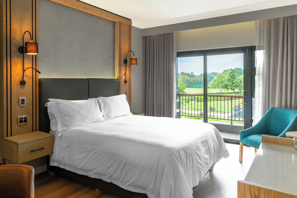 A hotel room with a large bed, a desk, a chair, and a view of a park through a large window.