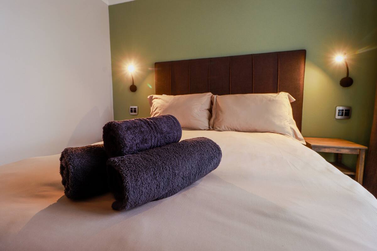 A neatly made bed with two rolled towels, a wooden headboard, and two wall-mounted lights.