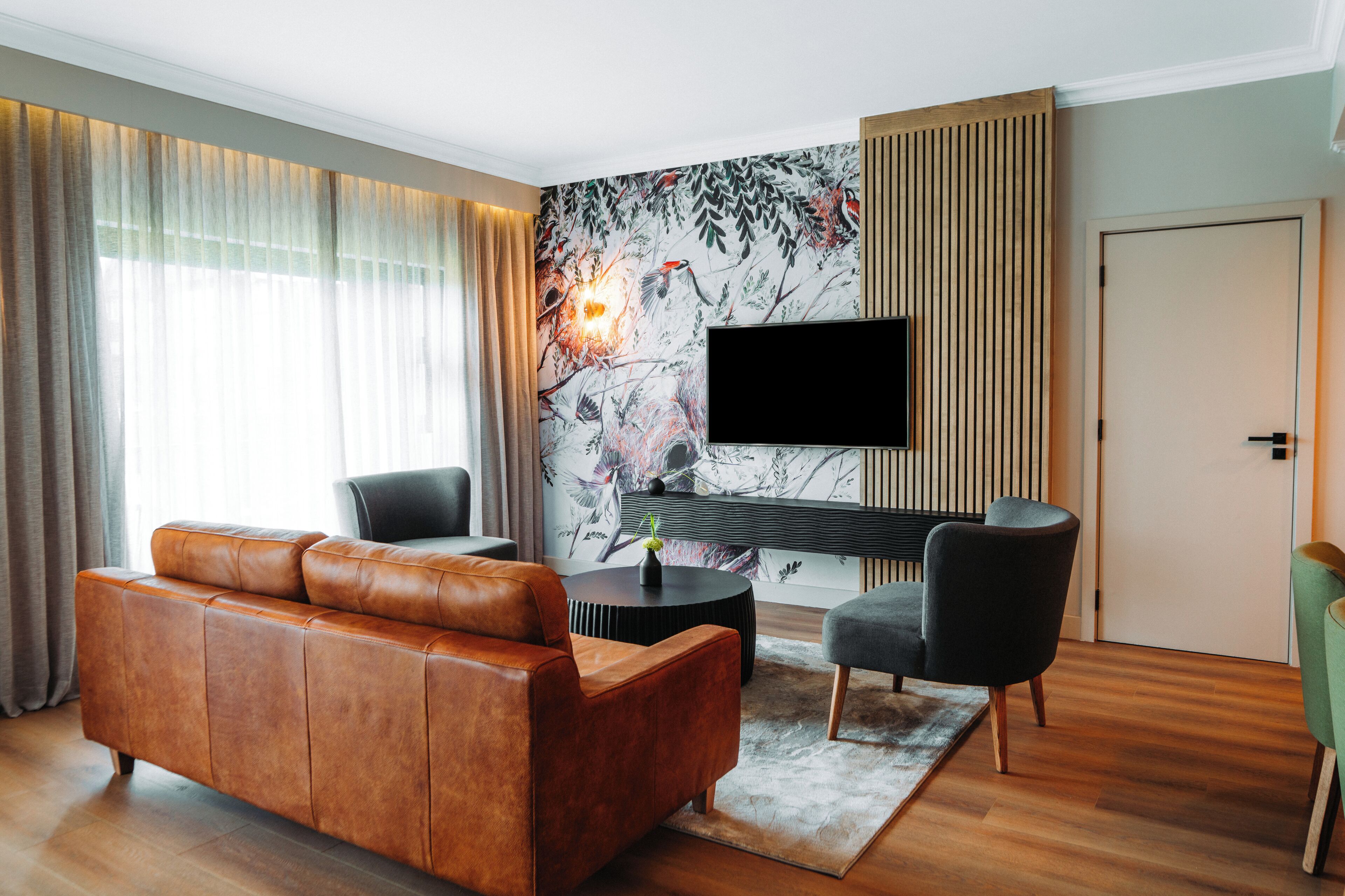 A modern living room with a leather sofa, two armchairs, a flat-screen TV, and a decorative wall mural.