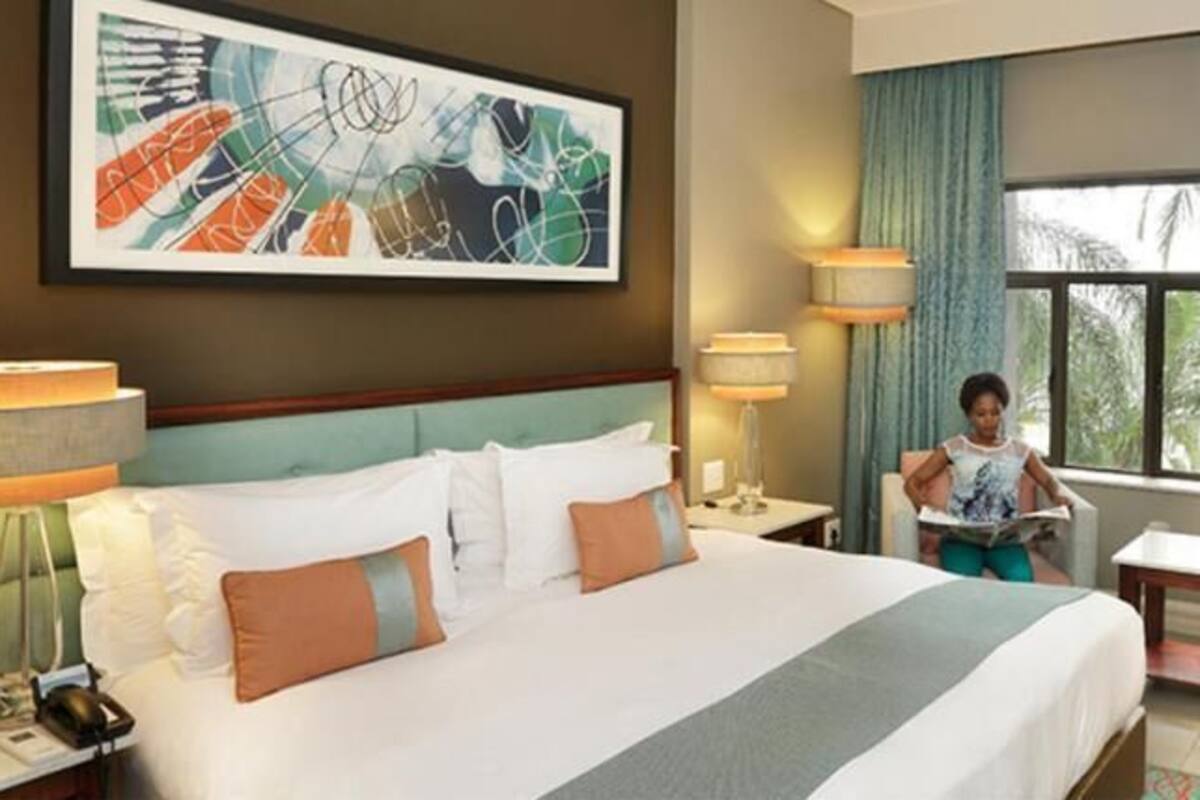 A hotel room with a bed, bedside tables, a desk, a chair, a window with curtains, and a painting on the wall.