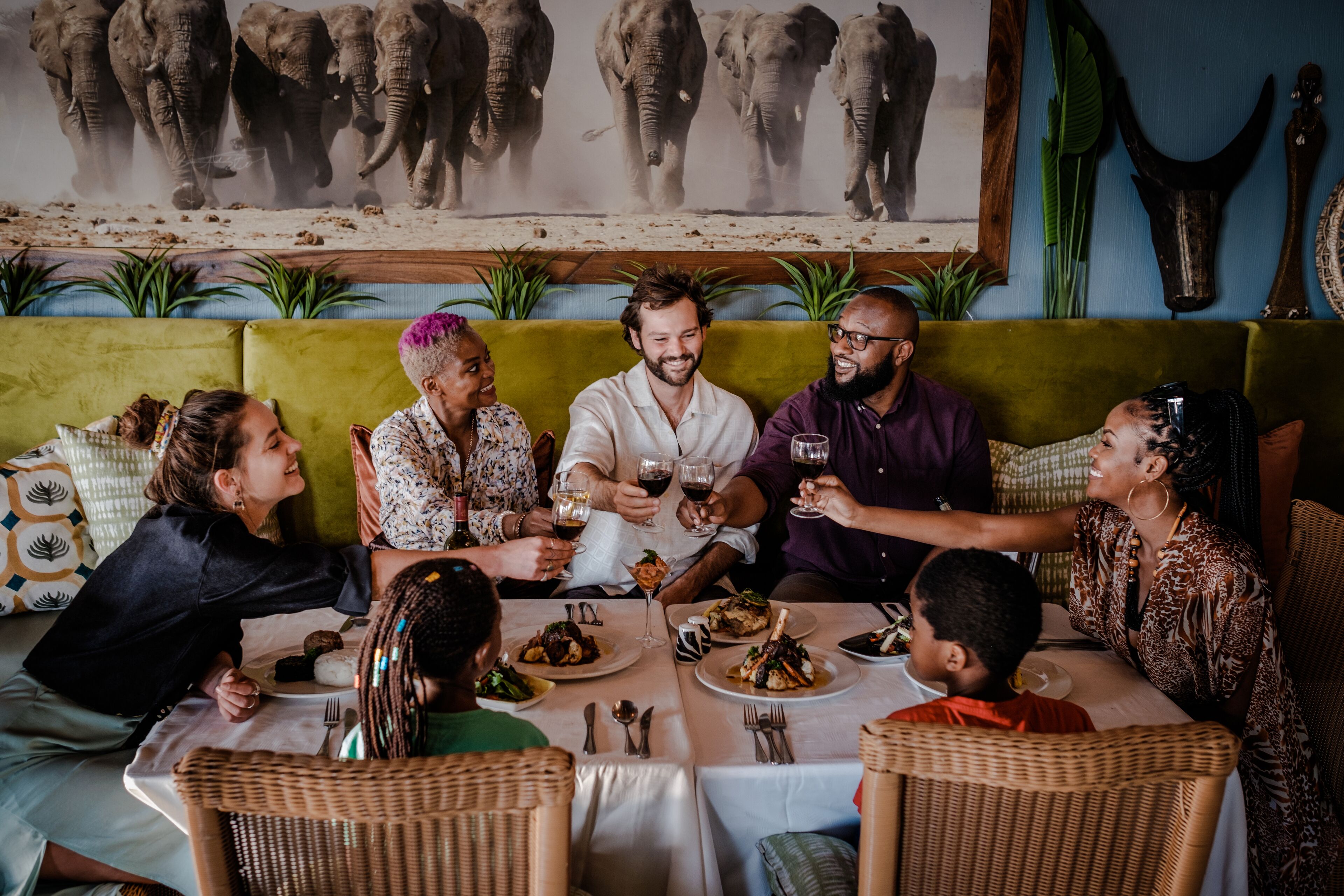 A group of people dining at a table with a large elephant painting in the background.